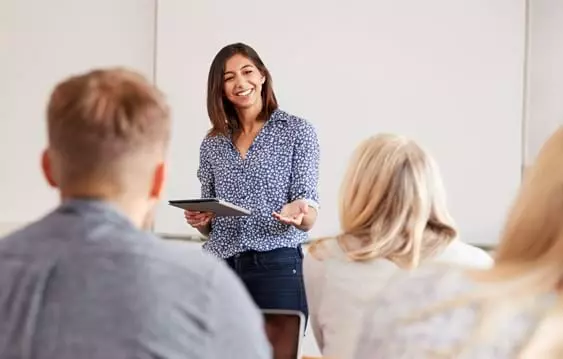 a smiling dark haired woman holding a notebook or tablet teaching a class