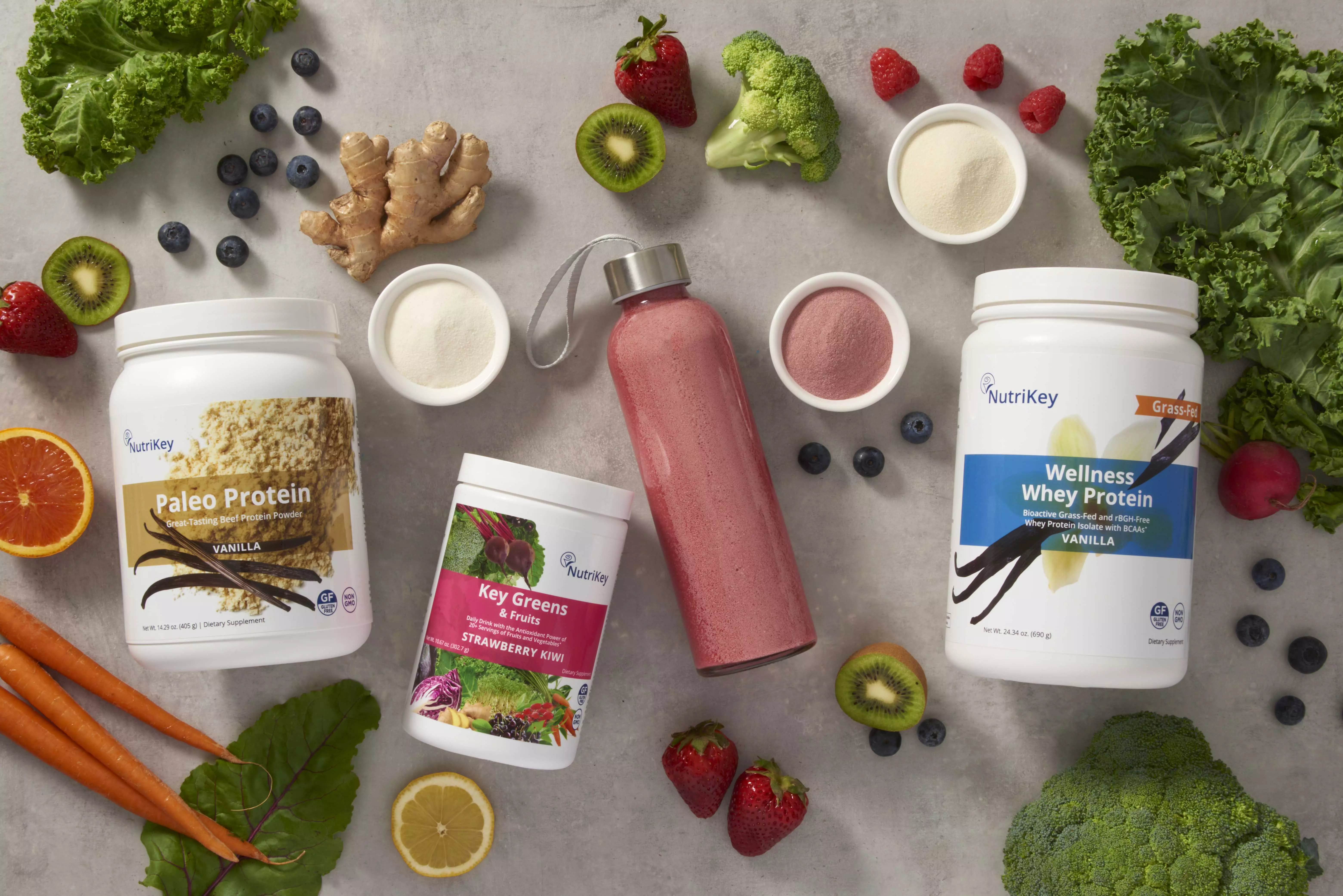 NutriKey protein powder and key greens products on a counter surrounded with various fruit and veggies