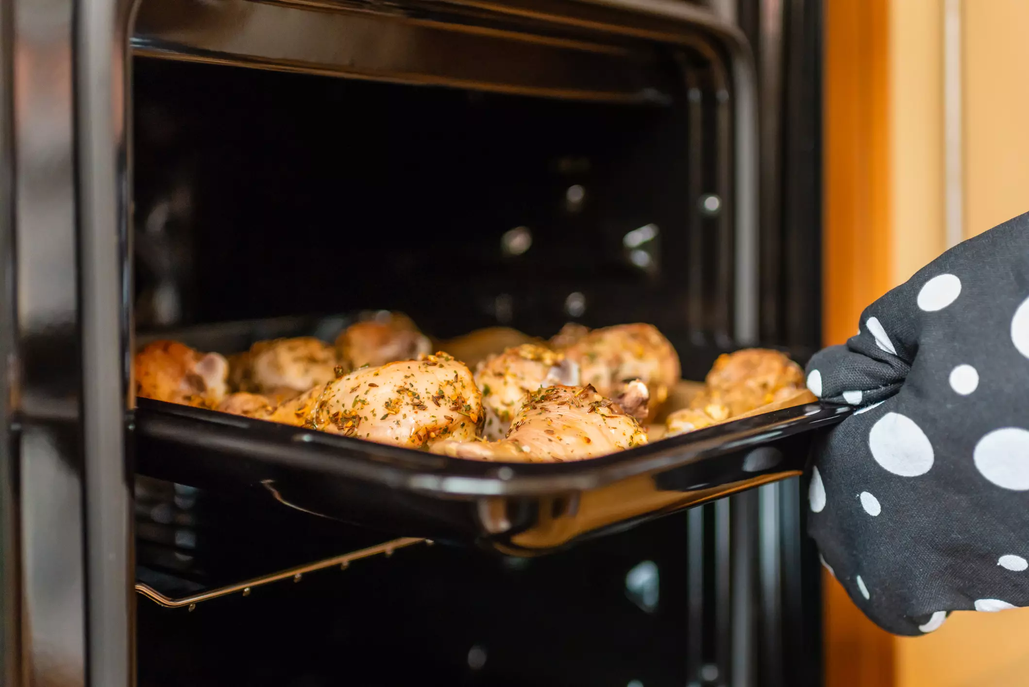 a hand in an oven mitt reaching taking out a sheet pan of chicken
