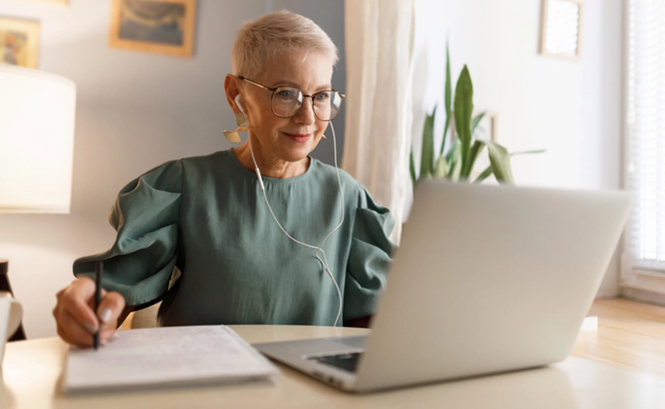 An older woman, with ear buds in, smiling at a laptop while prepared to take notes on a notepad